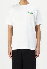 Bílý bavlněný tričko s krátkým rukávem, kulatým výstřihem a zelenožlutým potiskem "Na dovolené" na levém prsu. Černé kalhoty.