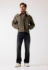 Veste courte vert olive avec col en shearling, boutons sur le devant, deux poches poitrine, associée à un jean foncé et des baskets blanches.