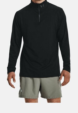 Mann trägt schwarzen Langarm-Quarter-Zip-Oberteil und hellgraue Shorts, steht mit entspannten Armen vor weißem Hintergrund.