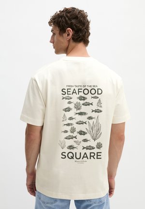 Mann trägt ein off-white T-Shirt mit schwarzem Meeresfrüchte-Motiv und Text auf dem Rücken, kombiniert mit hellblauen Jeans, mit dem Rücken zur Kamera.