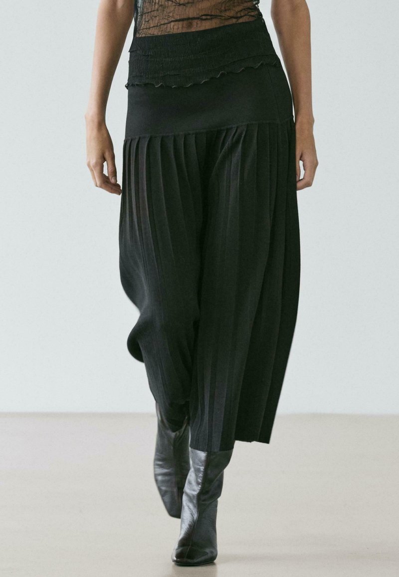 Pantaloni neri a gamba larga plissettati con una cintura testurizzata, caratterizzati da una finitura liscia e un tessuto fluido, abbinati a stivali neri alla caviglia.