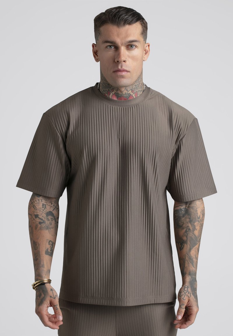 Homme tatoué portant une chemise marron à manches courtes côtelée et un pantalon assorti, se tenant avec une expression neutre devant un fond uni.