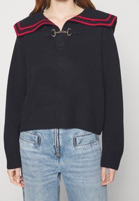 Marinblå stickad tröja med en rödrandig sjömanskrage, med en detaljerad guldlåsning och ribbad nederkant, kombinerad med ljusa denimjeans.