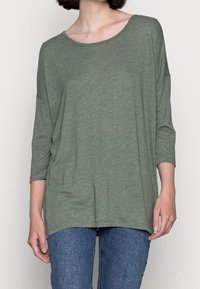 Vihreä, löysä yläosa, jossa on leveä kaula-aukko, 3/4-hihat ja teksturoitu, pehmeä kangas. Yhdistetty sinisiin farkkuihin.