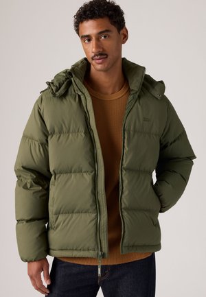 Homme aux cheveux courts et bouclés avec une moustache, portant une veste matelassée vert olive sur un pull marron, debout avec une main dans la poche.