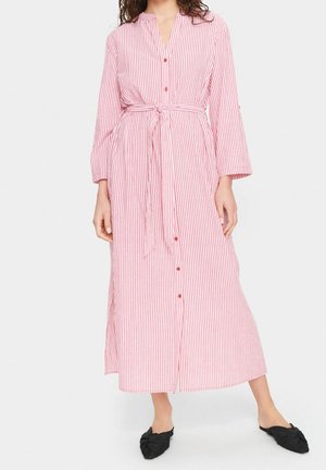 Kobieta ubrana w długą różowo-białą pionowo paskowaną sukienkę na guziki z przewiązaną talią i czarne czółenka na płaskim obcasie z ostrym noskiem, stojąca na białym tle.