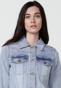 Svetlo modra jeans jakna z okrasnimi bleščicami, dvema žepoma na prsih in tradicionalnim ovratnikom. Ima sproščen kroj in gumbasto zapiranje.