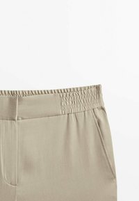 Beżowe spodnie z elastycznym paskiem i boczną kieszenią, gładki materiał i proste detale przeszyć.