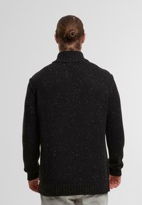 Schwarzer Strickpullover mit hohem Kragen, der ein strukturiertes Muster und weiße Spritzer aufweist. Ribbelbündchen und Saum.