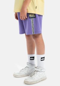 Paars shorts met een gedessineerde zijstreep, gedragen met witte hoge sneakers en witte sokken met een zwart logo.
