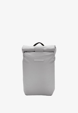 Sac à dos gris à rabat fabriqué en tissu durable, avec une poignée noire en haut et une zone d'étiquette à l'avant avec une texture lisse.