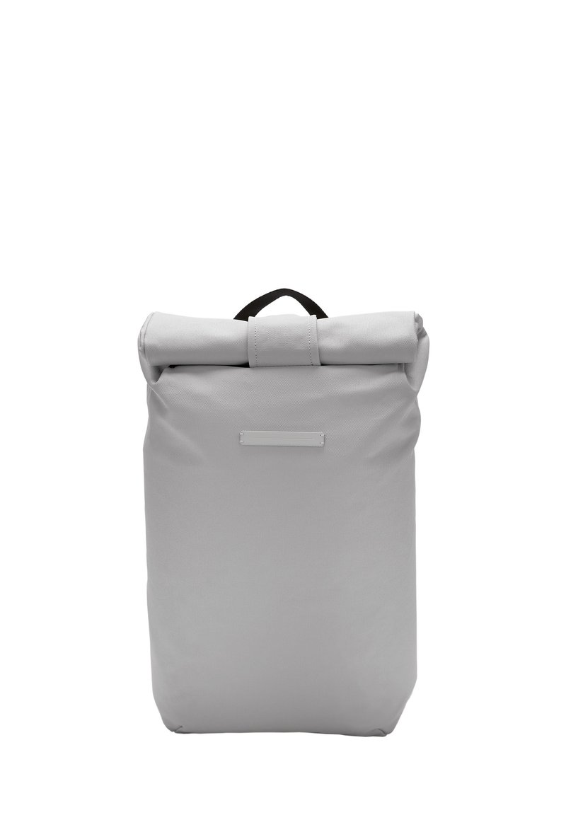 Sac à dos gris à rabat fabriqué en tissu durable, avec une poignée noire en haut et une zone d'étiquette à l'avant avec une texture lisse.