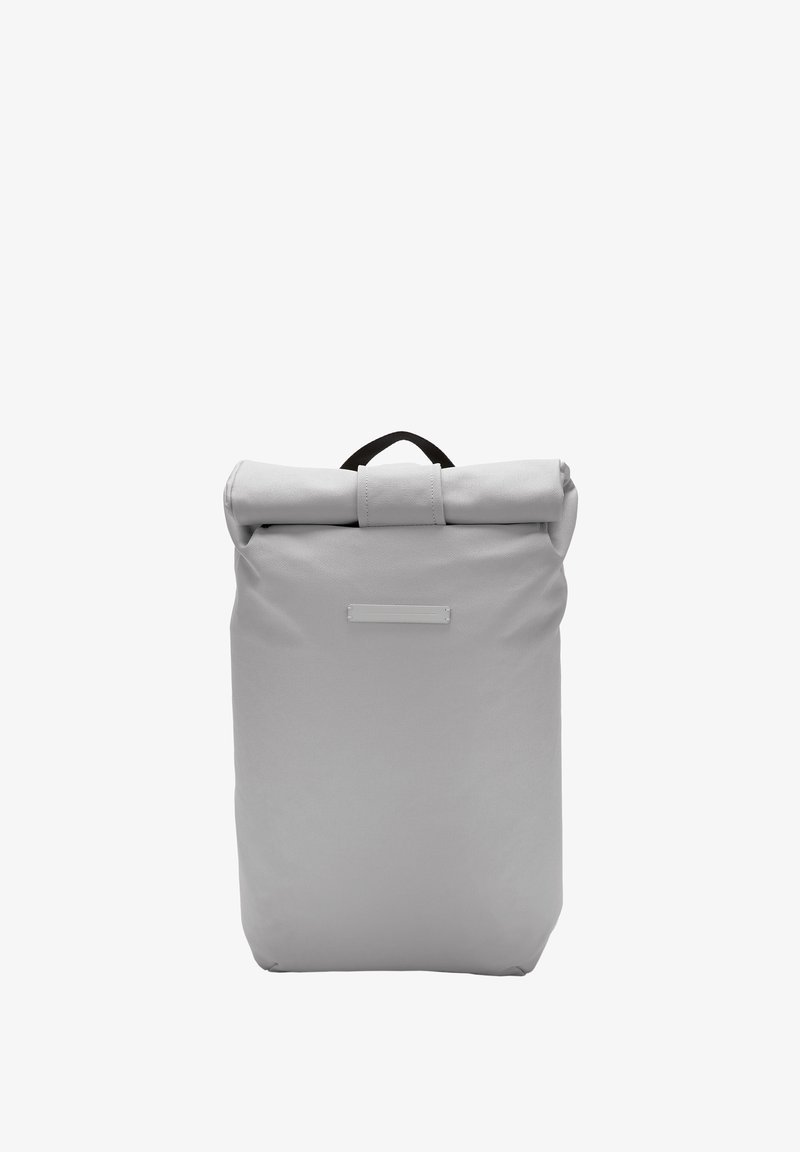 Sac à dos gris à rabat fabriqué en tissu durable, avec une poignée noire en haut et une zone d'étiquette à l'avant avec une texture lisse.