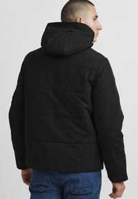 Sort polstret jakke med hætte, der har vandrette syninger for tekstur og en glat overflade. Bæres med blå denimjeans.