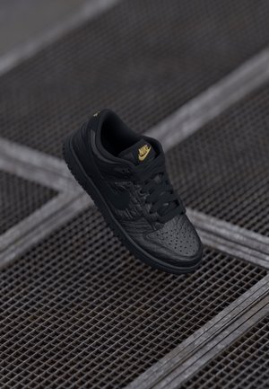 Zapatilla Nike de cuero negro con logo dorado flotando sobre un suelo de rejilla metálica en un entorno industrial tenuemente iluminado.