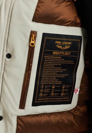 Poly ripstop pufferjack met een crèmekleurige buitenkant, bruine voering en gele rits. Voorzien van een zorglabel met specificaties, meerdere zakken en fleece details.