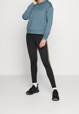 Personne portant un sweat à capuche bleu avec cordons, des leggings noirs à bandes jaunes sur les côtés, des chaussettes blanches et des chaussures de sport noires, debout devant un fond uni.