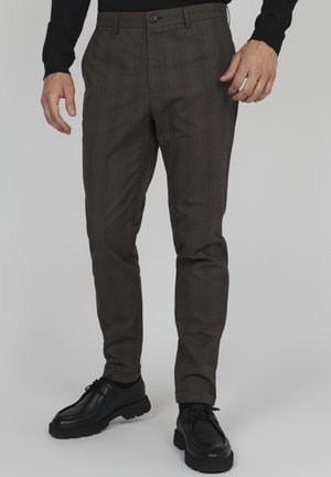 Mann trägt dunkelgraue schmal geschnittene Hose und schwarze Schnürschuhe, steht vor einem schlichten grauen Hintergrund.
