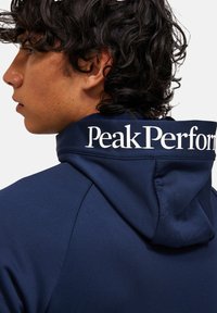 Tumedat sügisel hoodiga uppuvate kapuutsidega, mille taga on valge "Peak Performance" logo. Sile kangas minimaalsete õmblustega.