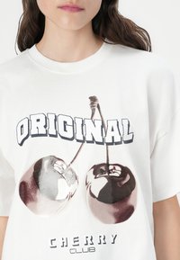Balta medvilninė marškinėlė su grafiniu spaudiniu, vaizduojančiu dvi vyšnias tamsiose ir šviesiose spalvose, viršuje esant užrašui „ORIGINAL CHERRY CLUB“. 