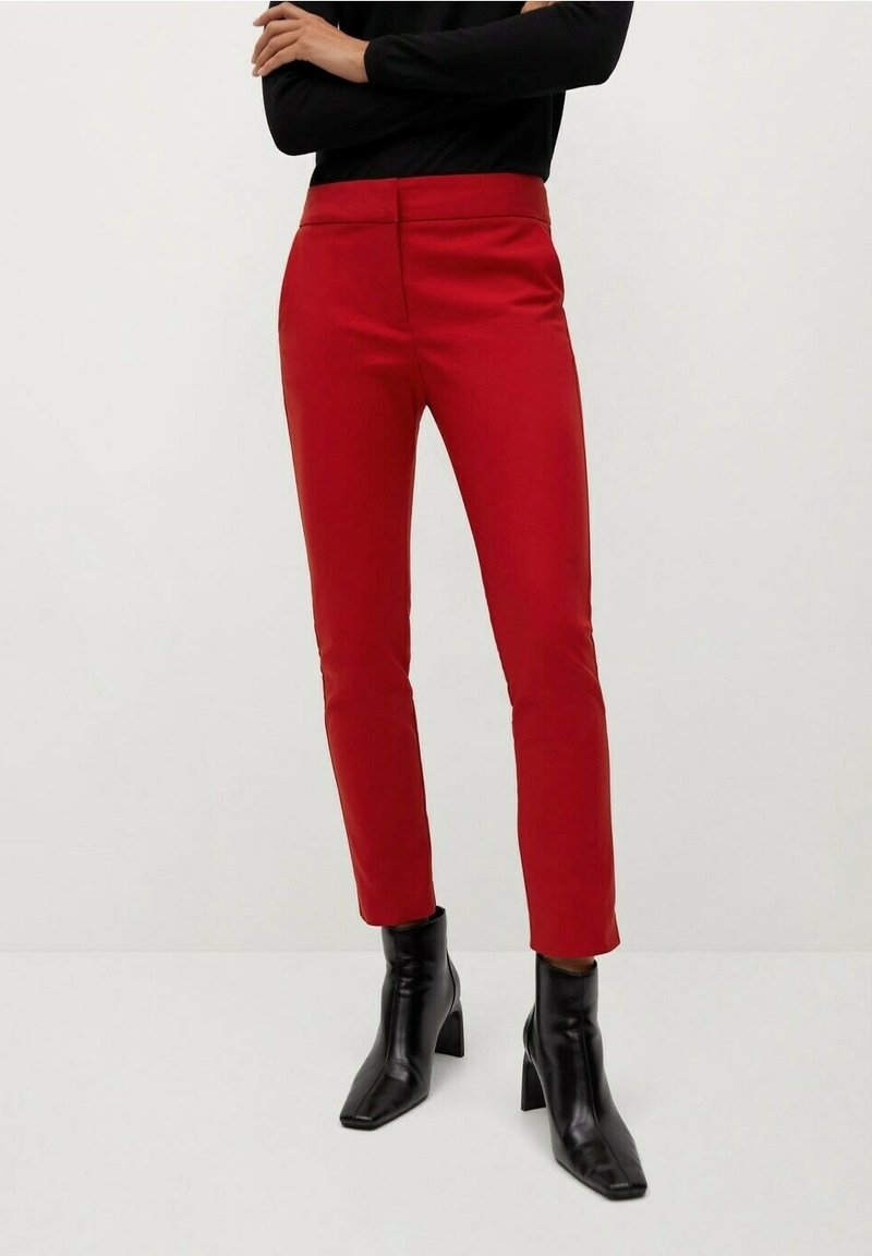 Personne portant un pantalon rouge slim et des bottines noires, debout les bras croisés sur un fond uni clair.