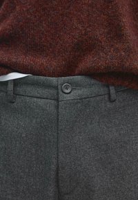 Gros plan d’une taille de pantalon grise texturée boutonnée associée à un pull en tricot couleur rouille et à un sous-vêtement blanc.