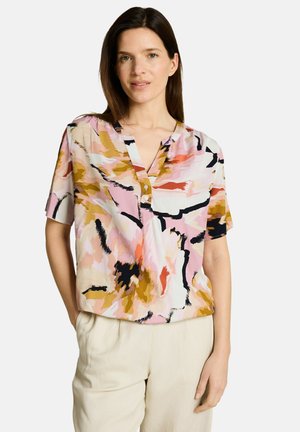 Frau trägt eine kurzärmelige Bluse mit einem abstrakten Muster in Pink, Beige, Schwarz und Orange und hellbeige Hose, steht mit einer Hand in der Tasche.