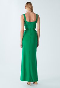 Sieviete ar gariem blondiem matiem, tērpusies zaļā bezrokās kleitā ar izgriezumiem viduklī un valkājot ādas krāsas augstpapēžu kurpes, stāv pie vienkārša fona.