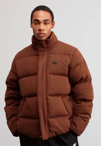 Giacca puffer marrone con colletto alto, caratterizzata da un logo Nike nero sul petto e un design squadrato. Tessuto liscio e imbottitura isolante.