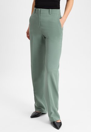 Hellgrüne, taillierte Hose mit geradem Schnitt, seitlichen Taschen und glatter Textur, kombiniert mit schwarzen spitzen Schuhen.
