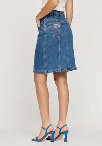 Jupe en denim bleu, longueur genou, avec poches arrière. Caractéristiques d'une coupe droite et d'un léger délavage. Portée avec des sandales à talons bleues.