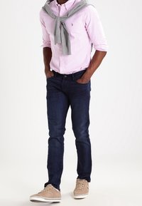 Jasnoróżowa, w paski, koszula z guzikami, szary sweter przewiązany na szyi, ciemnoniebieskie dżinsy i beżowe trampki z białą podeszwą.