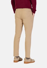 Pantalon chino beige, coupe fuselée, texture lisse, deux poches arrière, porté avec des chaussettes grises et des baskets beiges à semelle marron.