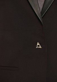 Sort blazer med satinrevers, der har et trekantet sølvmærke på venstre side og en paspoleret lomme på fronten.