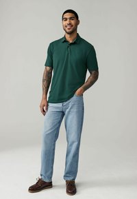 Dunkelgrünes Poloshirt mit klassischem Kragen, kombiniert mit hellblauen Jeans und braunen Leder-Schuhen, die lässigen Stil und Komfort zeigen.