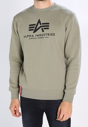 Mand iført olivengrøn Alpha Industries sweatshirt med sort logo og tekst, kombineret med mørke jeans på en ensfarvet lys baggrund.