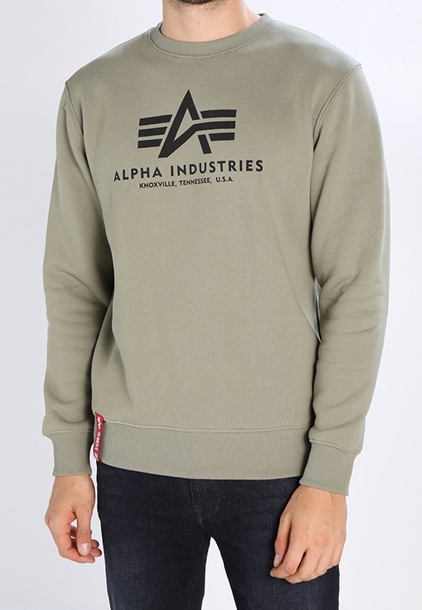 Homme portant un sweat-shirt vert olive Alpha Industries avec logo et texte noirs, associé à un jean foncé sur un fond clair uni.