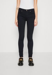 Jeans skinny in denim blu scuro, con vita a metà altezza, tasche frontali e dettagli in hardware dorato. Abbinati a mocassini neri.