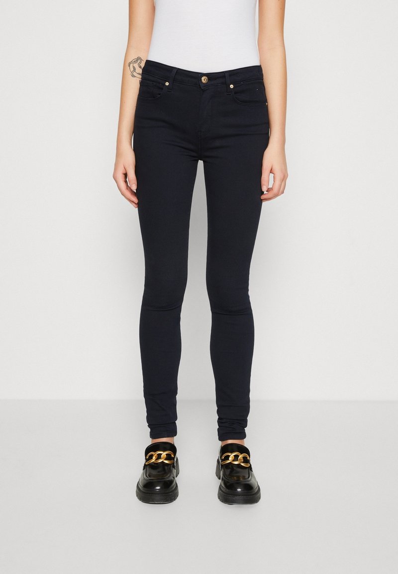 Jeans skinny in denim blu scuro, con vita a metà altezza, tasche frontali e dettagli in hardware dorato. Abbinati a mocassini neri.