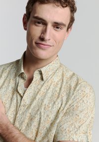 Helles kurzärmliges Hemd mit einem wirbelnden Blättermuster in Beigetönen und Hellblau, aus einem weichen, strukturierten Stoff gefertigt.