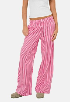 Kobieta ubrana w różowo-białe, pionowo paskowane spodnie o szerokich nogawkach z wiązanym w pasie sznurkiem oraz beżowe kapcie, stojąca na białym tle.