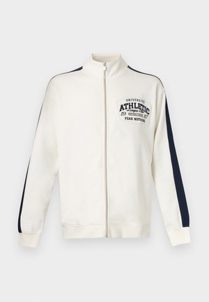 Veste de sport blanche zippée avec des rayures bleu marine le long des manches et le texte "University Athletic League Club 1987 Fear Nothing" sur la poitrine.