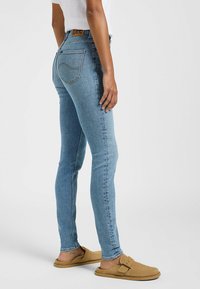 Jeans ajustados en denim azul claro. De tiro alto con bolsillos traseros, con una etiqueta marrón en la cintura. Textura suave con un desvanecimiento mínimo.