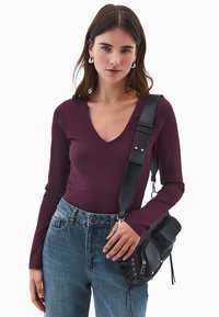 Giovane donna con capelli castani che indossa orecchini a cerchio argentati, un top viola a maniche lunghe a V, jeans blu e una borsa a tracolla nera con cinturino largo.