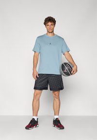 Lyseblå treningst-skjorte med svart logo, svarte mønstrede shorts og svarte/røde joggesko, holder en basketball med logodetalj.