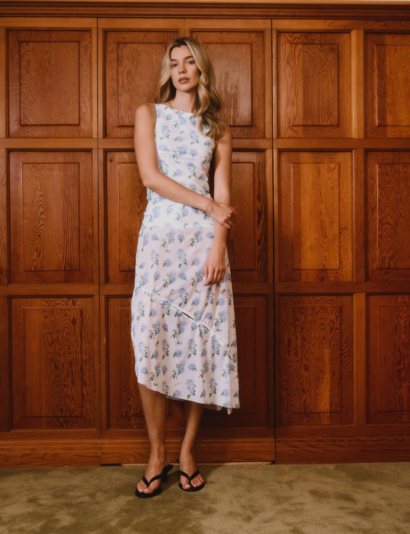 Γυναίκα με λευκό αμάνικο φόρεμα με μπλε λουλουδάτο σχέδιο στέκεται με τα χέρια σταυρωμένα απέναντι από ξύλινο τοίχο με πάνελ.