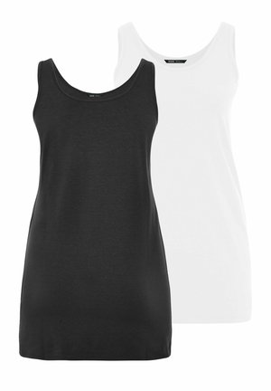 Lyshåret kvinde iført hvid ærmeløs tunika og blå jeans, stående mod en ensfarvet grå baggrund.