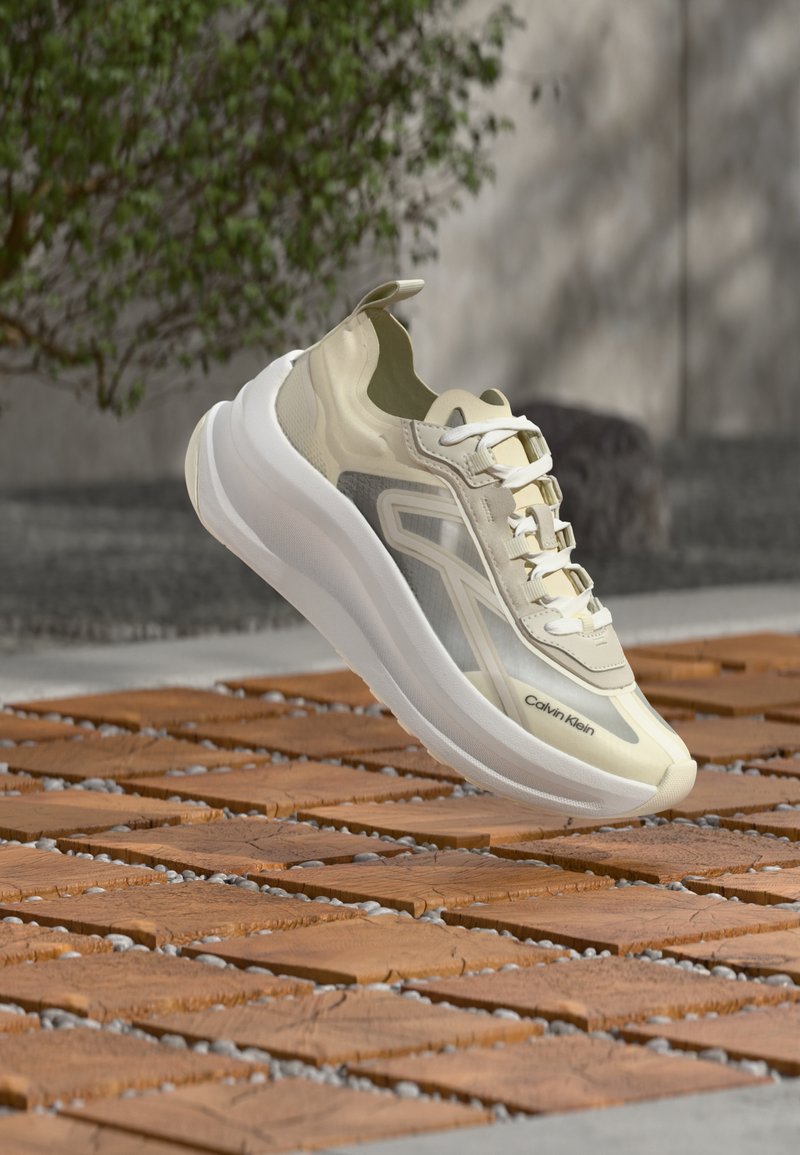 Μπεζ και λευκό αθλητικό παπούτσι Calvin Klein αιωρείται πάνω από ξύλινο πλακάκι σε εξωτερικό χώρο με φυτά και τοίχο σε απαλό φόντο.