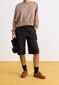 Brązowy krótki sweatshirt, czarne szorty ze sznurkiem, brązowe trampki, czarne skarpetki, trzymający czarną futrzaną torbę z białym napisem. Ustawione na żółtej podłodze.