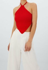 Haut dos nu rouge en tricot avec une forme triangulaire, doté de côtés ouverts et d'un nœud à l'arrière. Associé à un pantalon blanc taille haute.